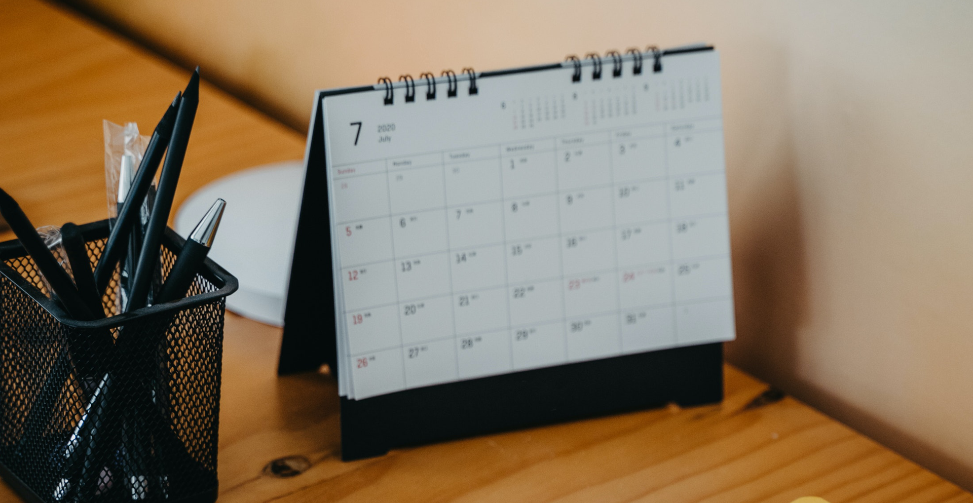 Zdjęcie przedstawia kalendarz stojący na biurku. Obok w pojemniku znajdują się przybory do pisania.