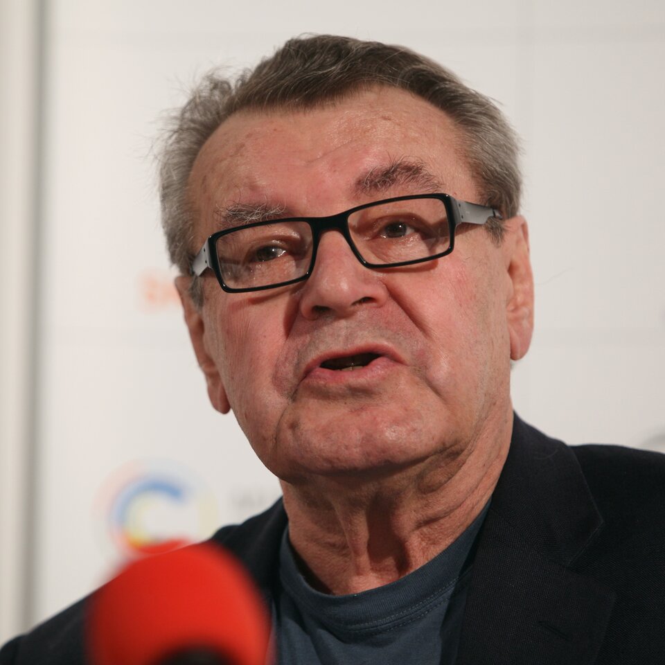 Zdjęcie przedstawia portret dojrzałego mężczyzny. Twarz mężczyzny jest owalna, widać zarysowane na niej zmarszczki. Mężczyzna ma krótko ostrzyżone, szpakowate włosy, nosi okulary. Ubrany jest w garnitur i podkoszulek. Mówi coś do stojącego przed nim mikrofonu.