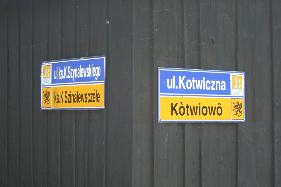 Fotografia przedstawiająca róg budynku. Na każdej ze ścian powieszone dwujęzyczne tablice z nazwami ulic. U góry lewej ściany znajduje się napis w języku polskim: ulica księdza K. Szynalewskiego obok umieszczony herb z błękitną literą J w kształcie rybackiego haczyka. Dodatkowym elementem jest wizerunek dwóch srebrnych ryb tworzących literę B. Poniżej napis w języku kaszubskim: księdza K. Szinalewsczéle obok umieszczony herb z czarnym gryfem na żółtym tle. To mityczne zwierzę z ciałem lwa oraz z głową i skrzydłami orła oraz uszami osła. Na głowie ma koronę, ma również otwarty dziób i wysunięty język. Na prawej ścianie tablice z napisami ulica Kotwiczna oraz Kòtwiowô.
