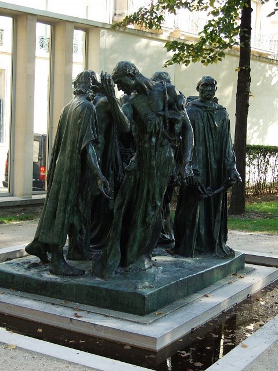 lustracja przedstawia rzeźbę pt. „Mieszczanie z Calais” autorstwa Augusta Rodina. Grupowa rzeźba ukazuje sześć odlanych w brązie postaci wielkości naturalnych. Postacie w luźnych szatach, stoją w rożnych pozach na tle budynku. Po prawej stronie widoczny jest pień drzewa i fragment gałęzi. Całość ma wymiary: 210 x 190 x 140 cm.