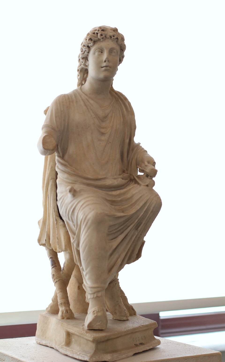 Jasna marmurowa rzeźba przedstawia siedzącą postać młodzieńca w długiej, miękko udrapowanej szacie. Figura ma kręcone włosy, spokojny wyraz twarzy i lekko uniesioną głowę zwróconą w bok. Jedna ręka jest ułamana w okolicy przedramienia, druga spoczywa na kolanie i trzyma zwinięty przedmiot. Postać siedzi na prostej podstawie, a tło jest jasne i neutralne, muzealne.