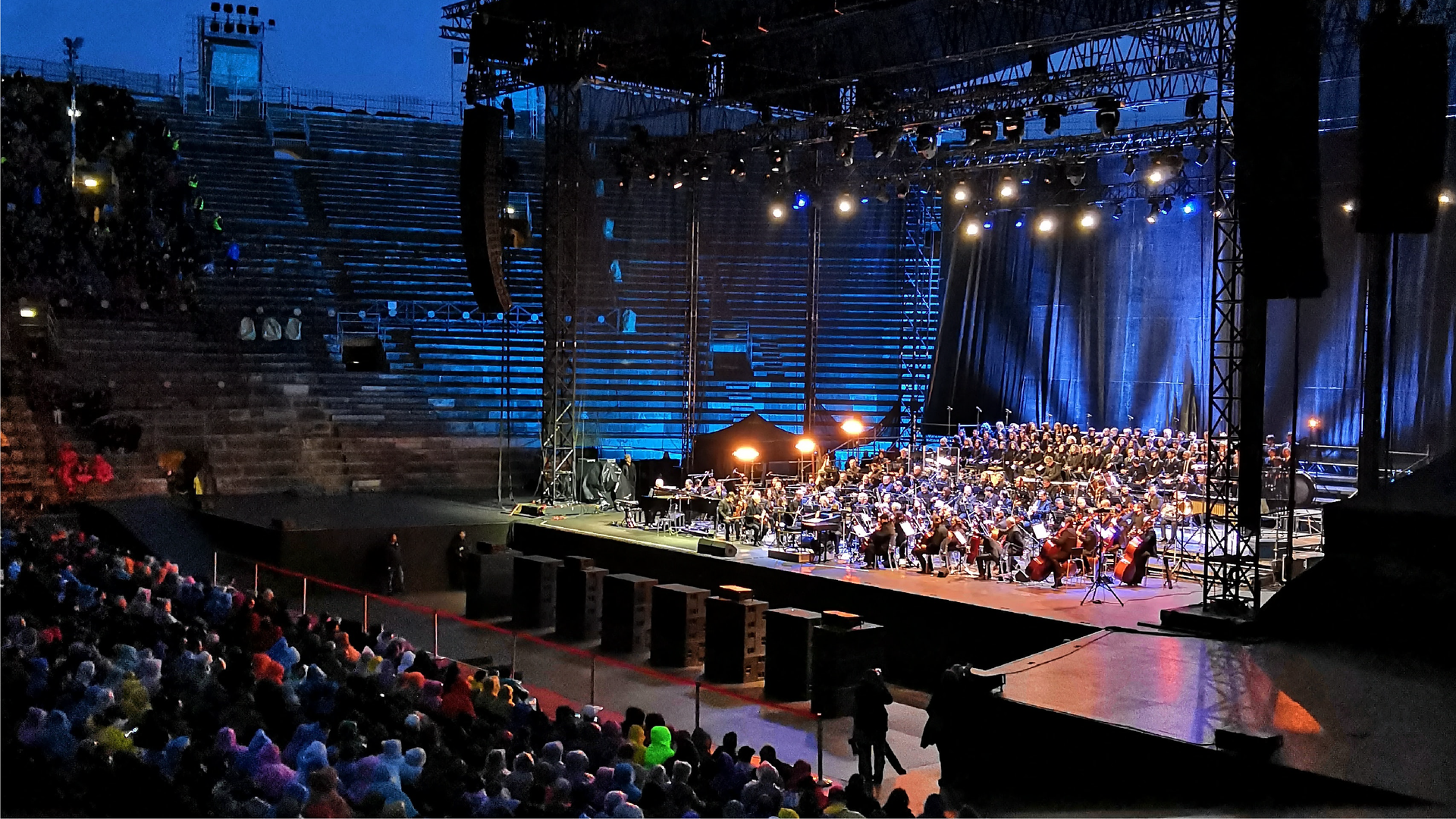 Zdjęcie przedstawia amfiteatr. Widoczna jest oświetlona scena, na której znajduje się orkiestra. Na wprost sceny siedzą w rzędach widzowie. W tle widoczne są także trybuny amfiteatru.