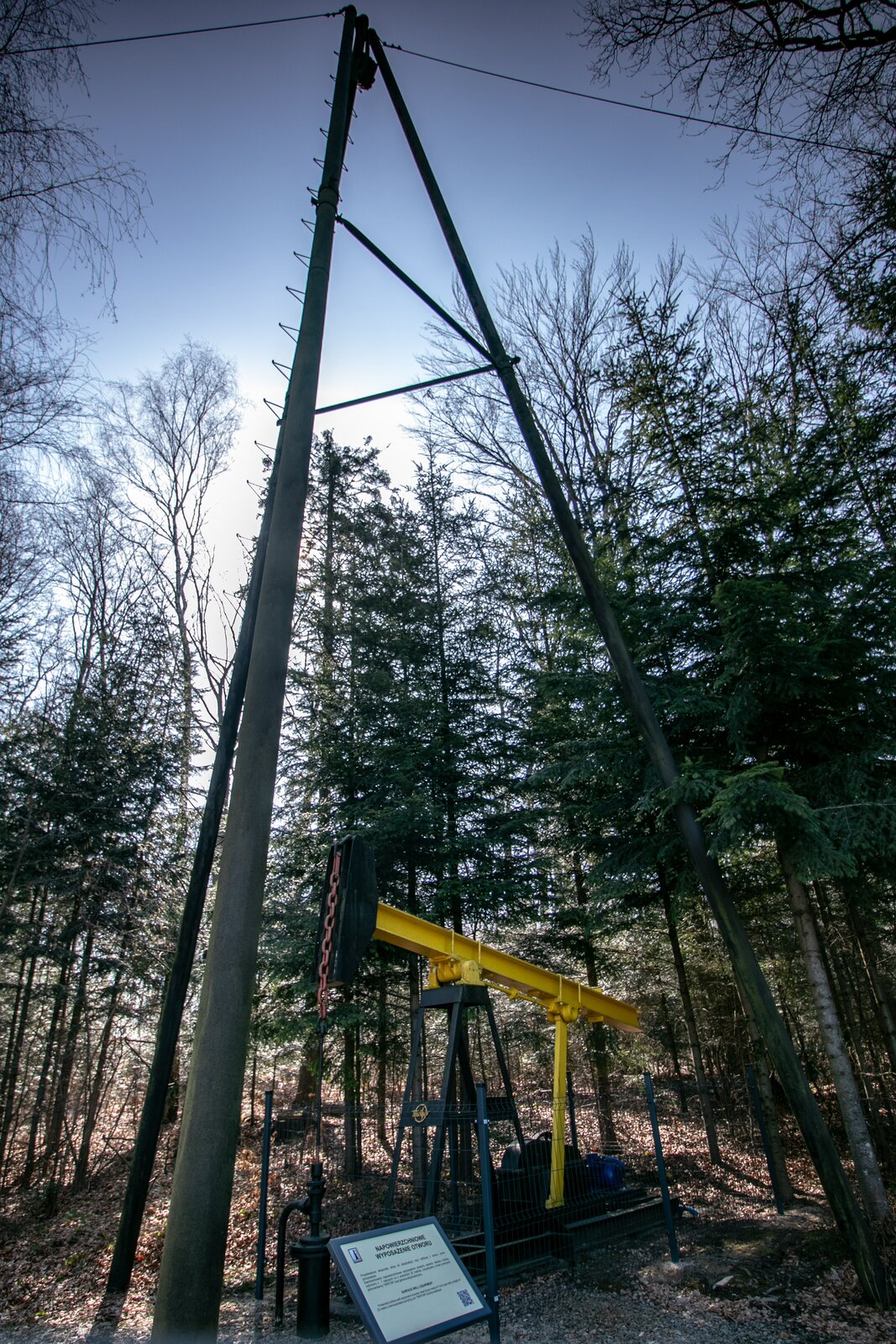 Na zdjęciu widać głowicę eksploatacyjną czyli urządzenie montowane na powierzchni terenu przy wylocie odwiertu, w celu jego zamknięcia oraz indywidualny żuraw pompowy. Urządzenia postawione jest w otoczeniu leśnym. Nad stalowym żurawiem pompowym, czyli urządzeniem wykorzystywanym do wydobycia surowca z górotworu rozstawiony jest trójnóg do obróbki odwiertu. Trójnóg jest kilkukrotnie wyższy od żurawia pompowego. Nogi trójnoga wykonane są z drewna. Żuraw pompowy umieszczony jest za siatką. Nad siatką wystaje stalowy wahacz wraz z czerwonym łbem. Element ten jest w kształcie litery T. Na płocie otaczającym żuraw pompowy znajduje się prostokątna tablica ostrzegawcza. Po lewej stronie żurawia znajduje się fragment rurociągu, który przyłączony do jego elementów. Drugi widoczny fragment rurociągu biegnie pod ziemię. 