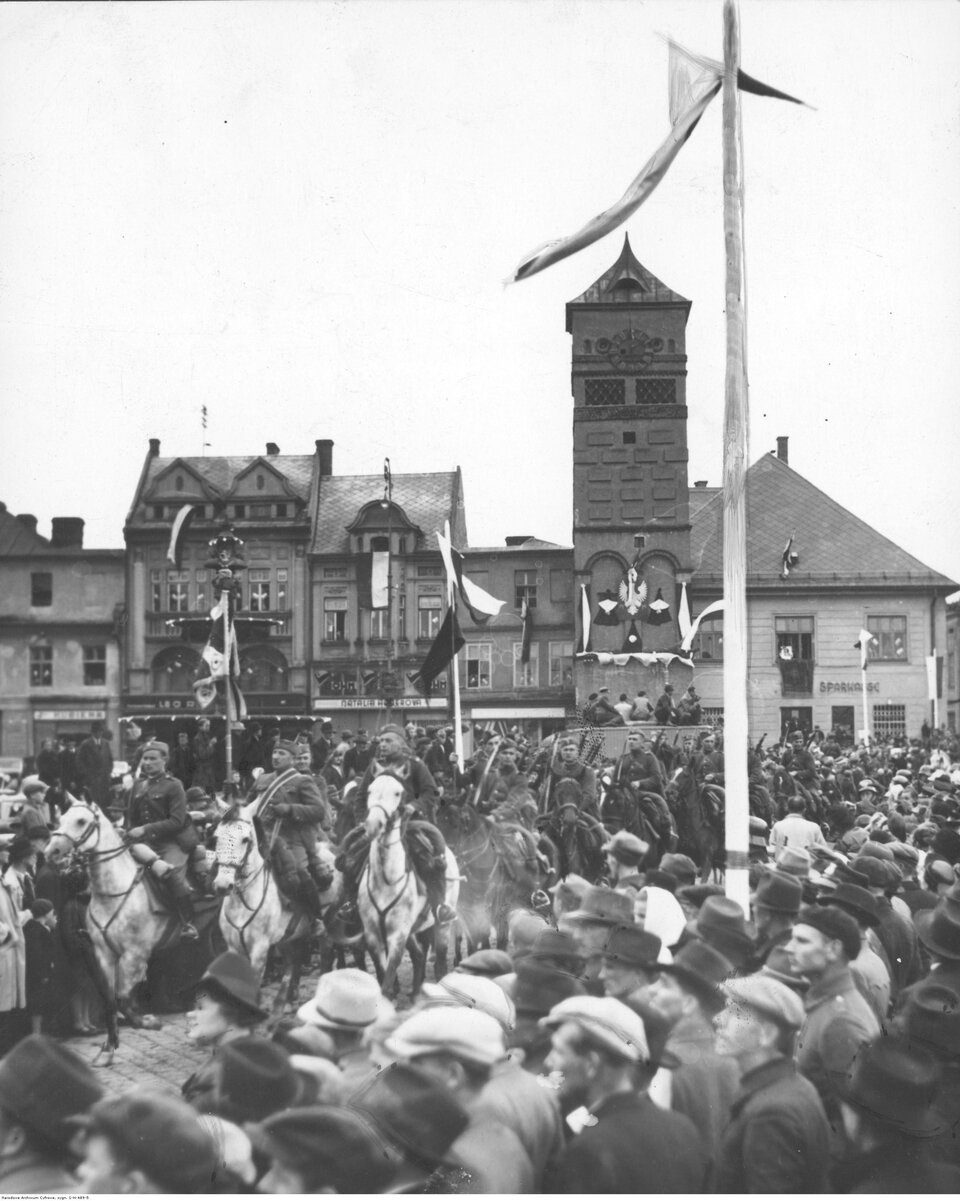 Zdjęcie przedstawia duży, miejski plac, po którym jedzie oddział kawalerii. Wzdłuż trasy przejazdu stoi tłum ludzi.