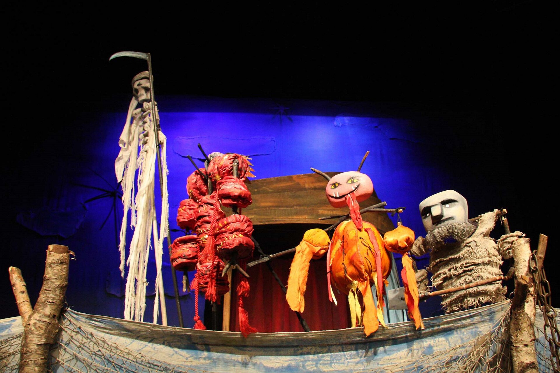 Ilustracja przedstawia kadr ze spektaklu „Ludowa szopka polska” Teatru Lalki Tęcza. Na fotografii widoczne są cztery drewniane lalki, które przedstawiają kolędników. Postacie są zwierzęcymi maszkarami. Po lewej stronie stoi kościotrup z kosą. Tło za szopką jest ciemnoniebieskie.