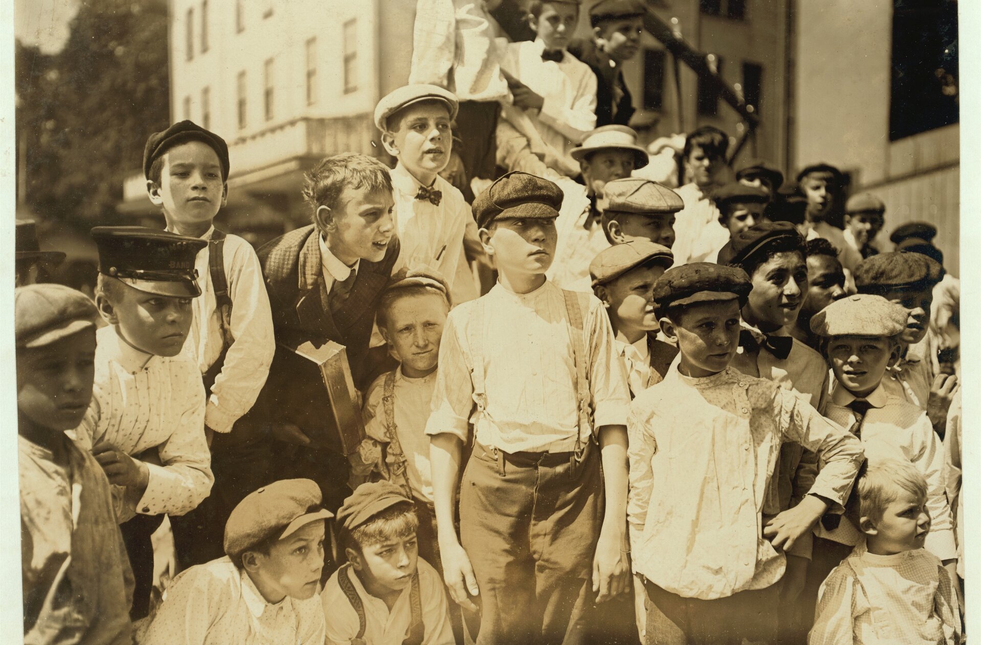 Zdjęcie przedstawia grupę chłopców w czapkach i koszulach. Dzieci stoją zgrupowane na ulicy. Patrzą się przed siebie.