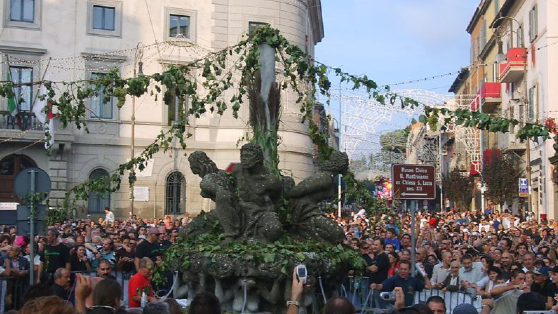 Zdjęcie przedstawia tłum ludzi na ulicy w czasie jakiegoś święta. W centralnym punkcie widoczny jest pomnik, od którego odchodzą girlandy zrobione z roślin. Wokoło widać włoskie kamienice.