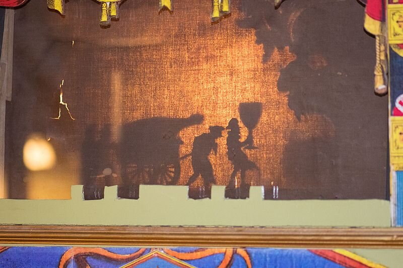 Zdjęcie przedstawia teatr cieni. Padający cień przedstawia kilka postaci oraz wóz.