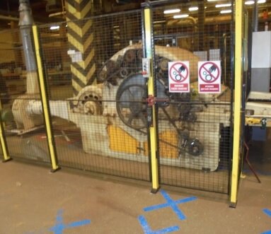 Zdjęcie przedstawia wilka zgrzeblącego - maszynę do produkcji wyrobów z włókna kokosowego. Stoi w hali zakładu produkcyjnego. Jest zabezpieczona ogrodzeniem z siatki. Na siatce znaki ostrzegawcze. Maszyna w środkowej części ma walcowaty kształt. W tylnej części połączona jest z rurą, skierowaną w stronę sufitu.
