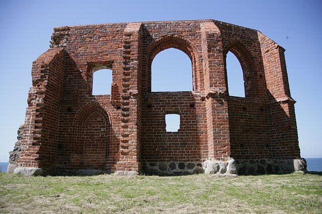 Na fotografii ruiny kościoła w Trzęsaczu. Po budowli pozostała jedynie kilkumetrowej długości ceglana ściana z czterema otworami okiennymi oddzielonymi od siebie pozostałościami muru. Przez okna w perspektywie widać niebo, a z boku ruin fragment morza. 