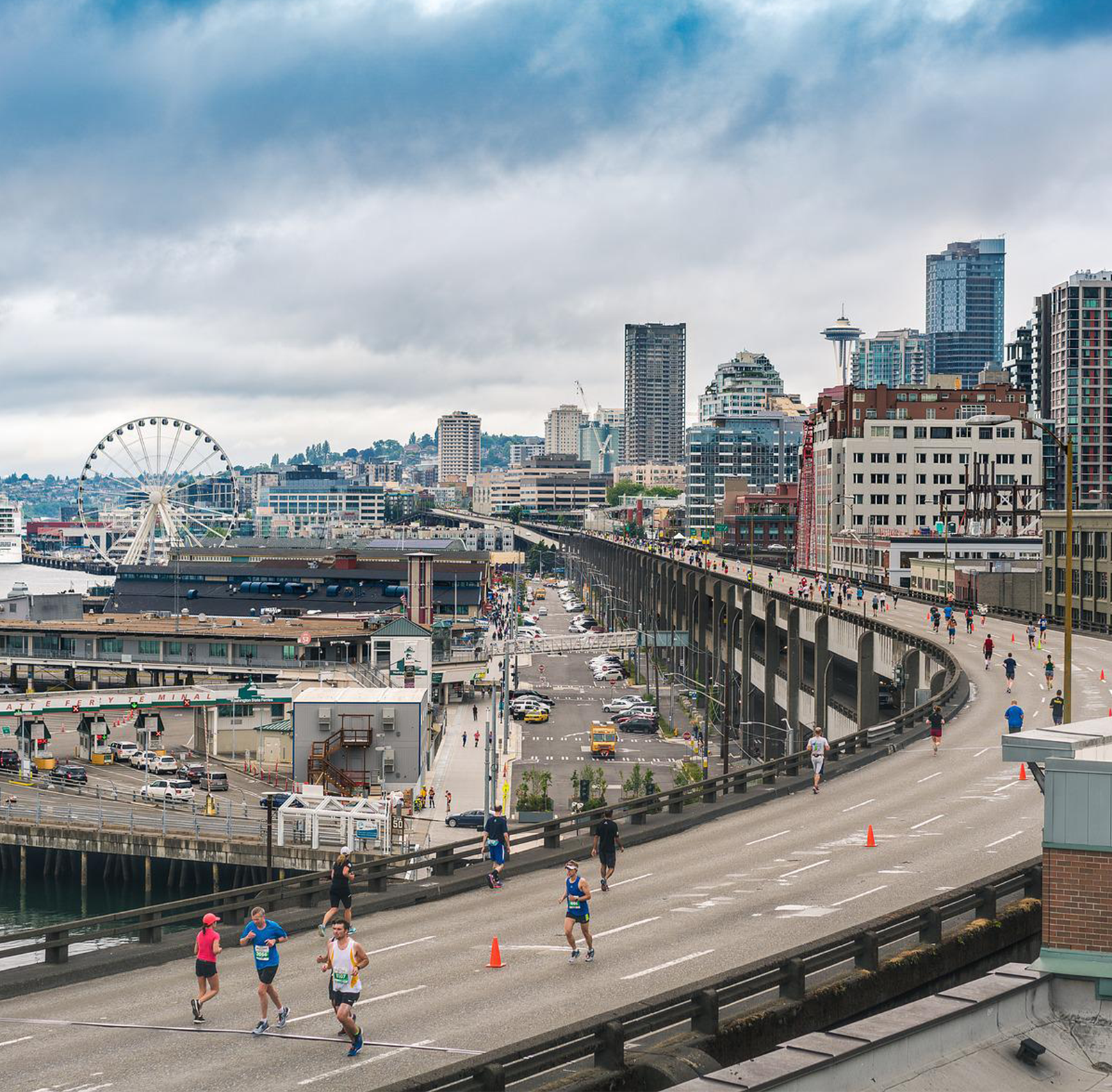 Zdjęcie przedstawia ulicę biegnącą w mieście, na której odbywa się maraton. Widoczne są budynki wielokondygnacyjne, parking oraz karuzela koło i ludzie biegnący w maratonie. 