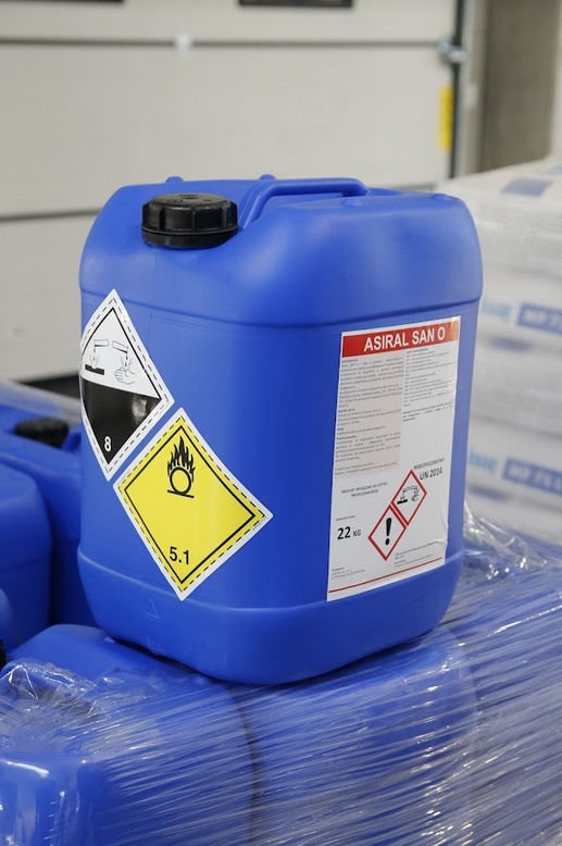 Zdjęcie przedstawia plastikowy kanister z oznaczeniami. Jedno z nich jest rombem z górną połową w kolorze białym i dolną w czarnym. Oznaczenie posiada cienką czarną obwódkę. W górnej części znajduje się rysunek dwóch probówek, z których wylewa się ciecz na powierzchnię płaską i na ludzką dłoń, które obie parują. Obok Znajduje oznaczenie będące żółtym rombem z czarną cienką obwódką. W górnej części rombu czarny płomień, a pod nim krótki poziomy odcinek.