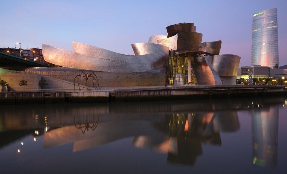 Fotografia przedstawia budynek położony nad  rzeką, na tle ciemniejącego nieba oraz innych budynków. Ściana z wejściem do budynku jest wykonana z połyskującego metalu. Przy wejściu do budynku znajduje się gruby, kremowy filar. Podtrzymuje on konstrukcję wysuniętego dachu, którego fragmenty przypominają srebrne kadłuby łodzi. Ściany również pozbawione są kątów prostych, a ich kształt także przywodzi na myśl długie łodzie. Przed budynkiem uchwycony jest brukowany plac, na którym umieszczono rzeźbę – dużego pająka na cienkich, wysokich nogach. 