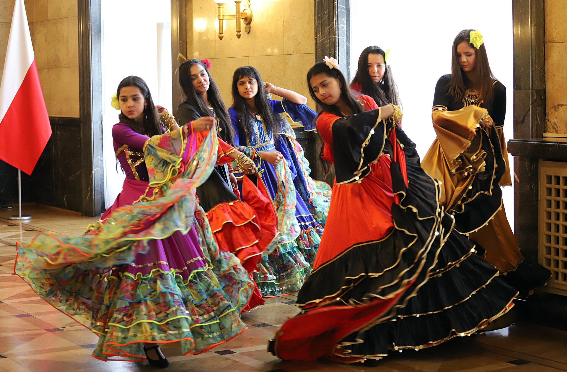 Zdjęcie przedstawia taniec, w wykonaniu sześciu kobiet. Stroje tańczących kobiet charakteryzują się bogatą kolorystyką i nasyconymi barwami. Każda z kobiet ma wpięty we włosy kwiat. Taniec odbywa się w sali, w której po lewej stronie umiejscowiona jest biało‑czerwona flaga.