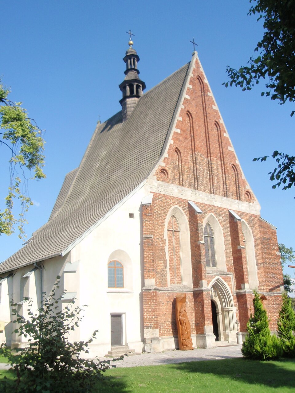 Fotografia przedstawia ceglany budynek w kształcie trójkąta ze stromym dachem. Pośrodku dachu widać wieżę z dzwonem w środku i krzyżem na czubku. Drzwi i okna budynku mają okrągłe sklepienia. Obok wejścia stoi drewniana figura. Wokół rosną drzewa. 