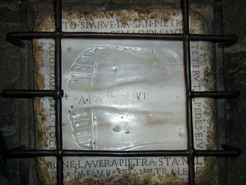 Zdjęcie przedstawia odciśnięte w kamieniu ślady stóp. Wokół odcisku stóp widać łacińskie napisy. Na kamień z odciskiem stóp nałożona jest żelazna krata z dużym otworem na ten odcisk.