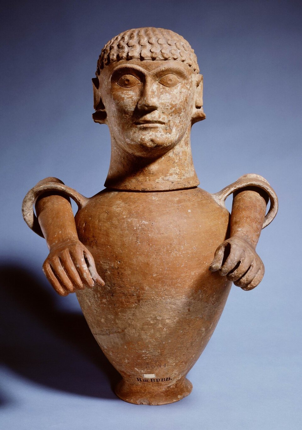 Ilustracja przedstawia urnę - jej kształt przypomina ludzką postać. Ręce postaci są modelowane jako część uchwytów urny. Pokrywa urny została uformowana w kształcie głowy mężczyzny o surowym wyrazie twarzy. 