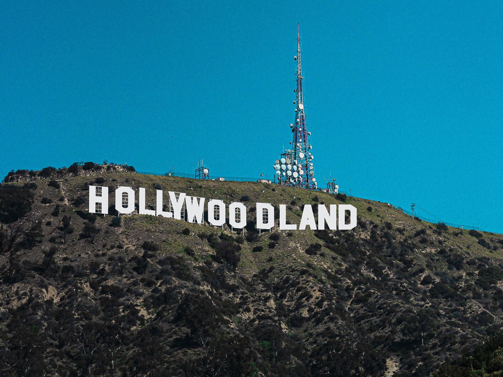 Zdjęcie przedstawia wzgórze z napisem z dużych białych liter w języku angielskim: HOLLYWOODLAND. Na szczycie widoczny jest maszt przekaźnika, na którym widoczne są anteny satelitarne. 
