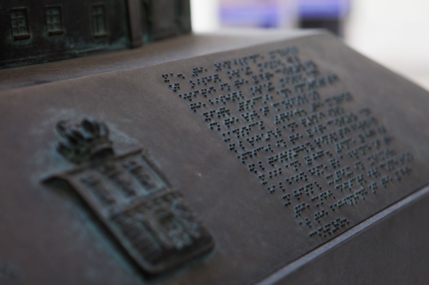Zdjęcie przedstawiające zapis alfabetem Braille'a [czytaj: brajla] umieszczony przy ekspozycji. Na metalowej płycie, z lewej strony znajduje się płaskorzeźba herbu, a z prawej stronie umieszczono zapis w alfabecie Braille'a [czytaj: brajla]. 