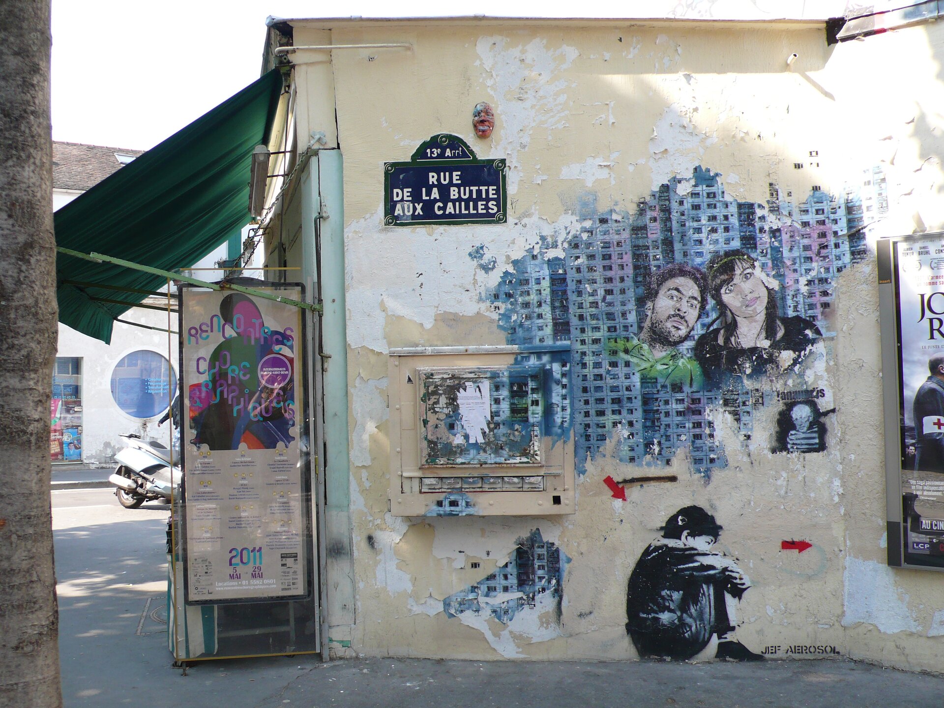 Ilustracja przedstawia boczną ścianę jakiegoś budynku, na którym narysowane jest graffiti z kobietą i mężczyzną na tle bloku mieszkalnego. Poniżej, na samym dole jest rysunek dziecka siedzącego na podłodze, opierającego głowę o kolana. Na budynku znajduje się tabliczka z napisem: RUE DE LA BUTTE AUX CAILLES.