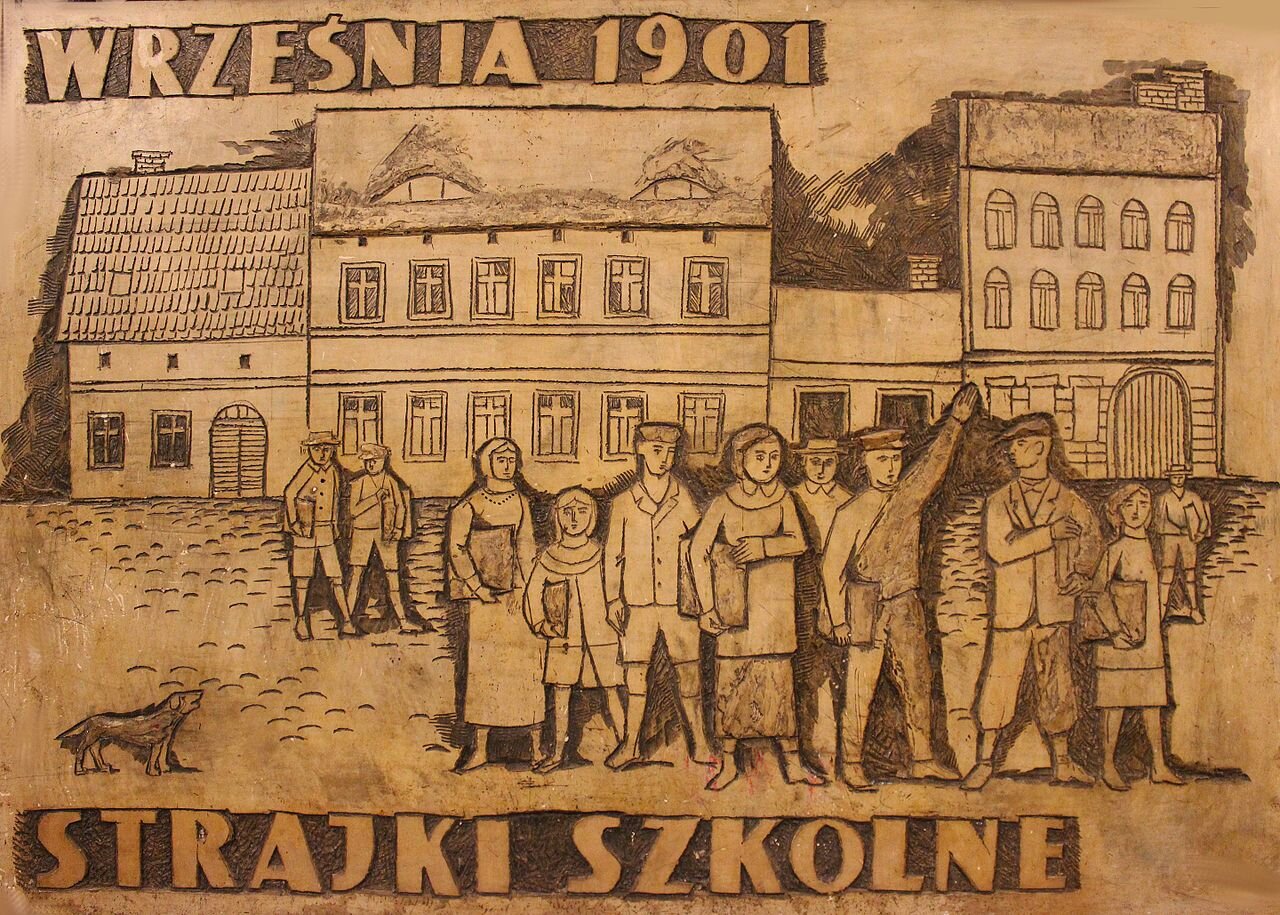 Zdjęcie przedstawia płaskorzeźbę. Prezentuje ona dzieci, które trzymają w rękach teczki. Dziewczynki mają sukienki i chusty na głowach, a chłopcy mundurki z czapkami. Jeden z chłopców unosi rękę. Za postaciami stoi rząd kamienic. Na górze jest napis: Września 1901. Na dole jest napis: Strajki szkolne.