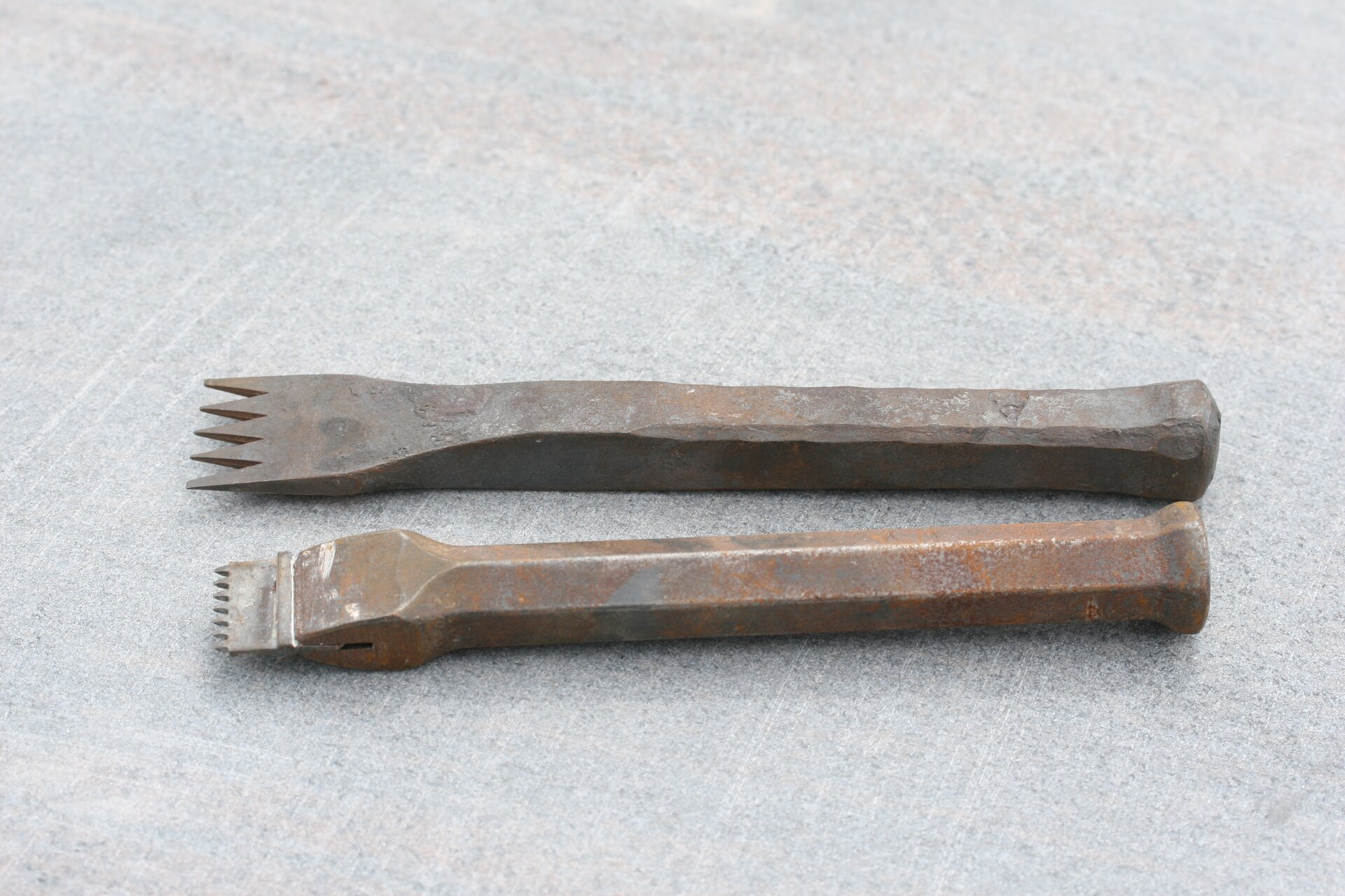 Fotografia przedstawia dwa metalowe przecinaki kamieniarskie. Oba są metalowe. Ten przedstawiony u góry ma bardziej prostokątną rękojeść, jest ciemniejszy oraz swoim kształtem przypomina widelec o drobnych, trójkątnych ząbkach. Drugi natomiast jest w kolorze miedzi i składa się z rękojeści oraz przytwierdzonego do niej metalowego elementu z bardzo małymi ząbkami.