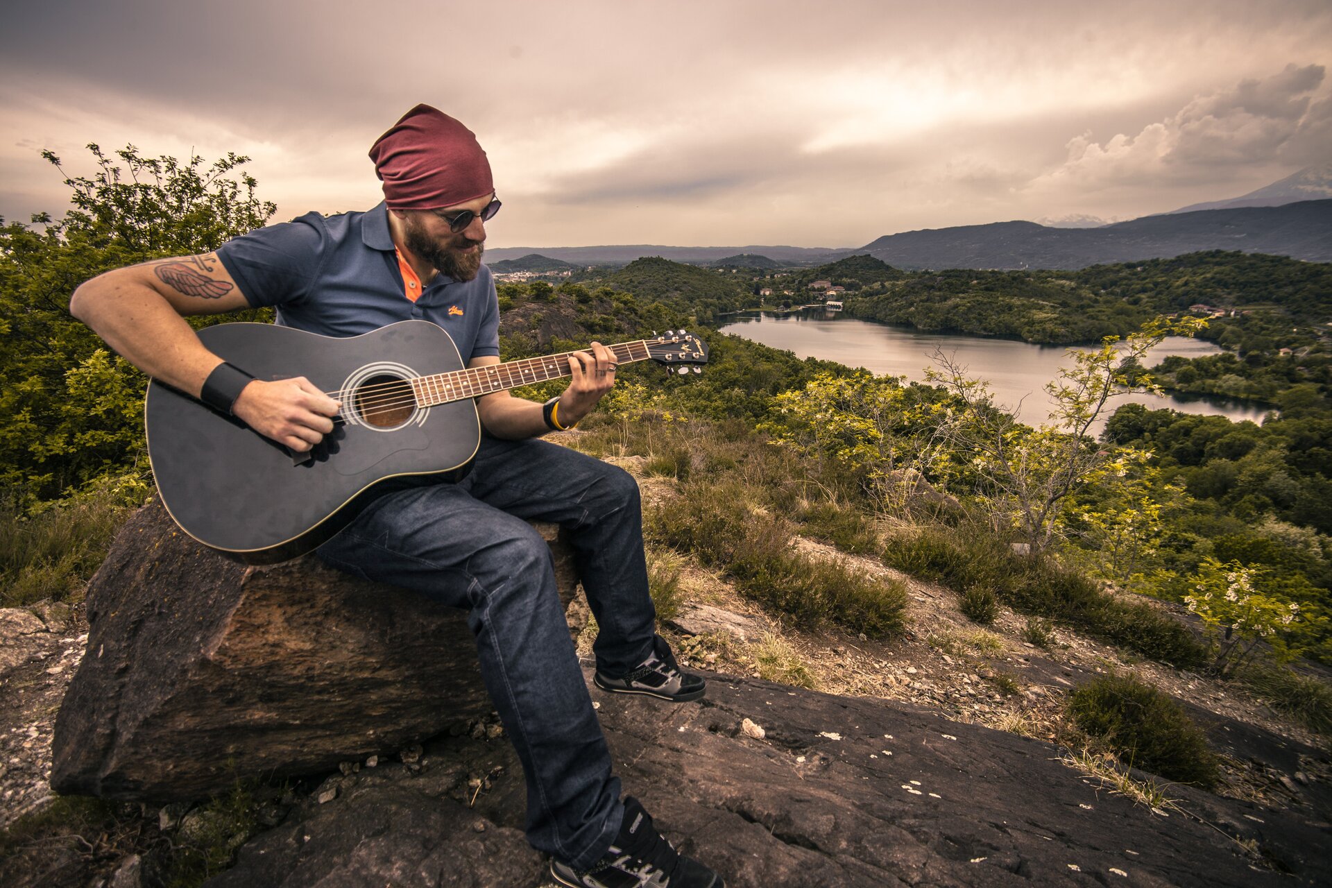 Zdjęcie przedstawia mężczyznę, który siedzi na skale i gra na gitarze. Mężczyzna ma ciemną brodę, założone okulary przeciwsłoneczne oraz czerwoną chustę na głowie. Ubrany jest w niebieską koszulką i dżinsy. W tle znajdują się lasy, jezioro oraz pasmo górskie.