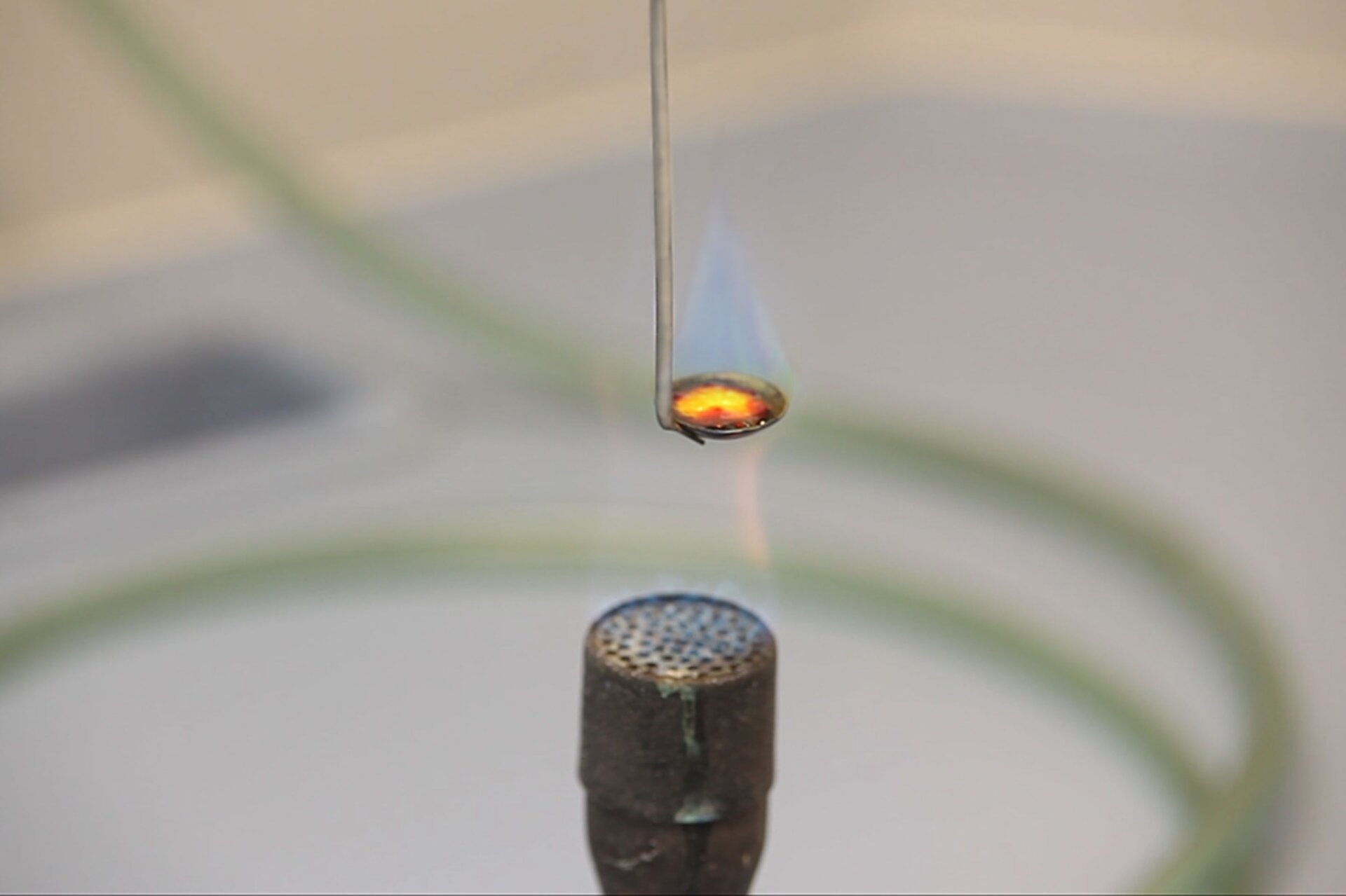 Zdjęcie przedstawia końcówkę palnika w zbliżeniu. Nad nim, w zasięgu słabo widocznego płomienia wisi laboratoryjna łyżeczka do spaleń, na której znajduje się paląca się siarka. Z łyżeczki unosi się biały dym, co jest jednym z elementów charakterystycznych reakcji spalania siarki.