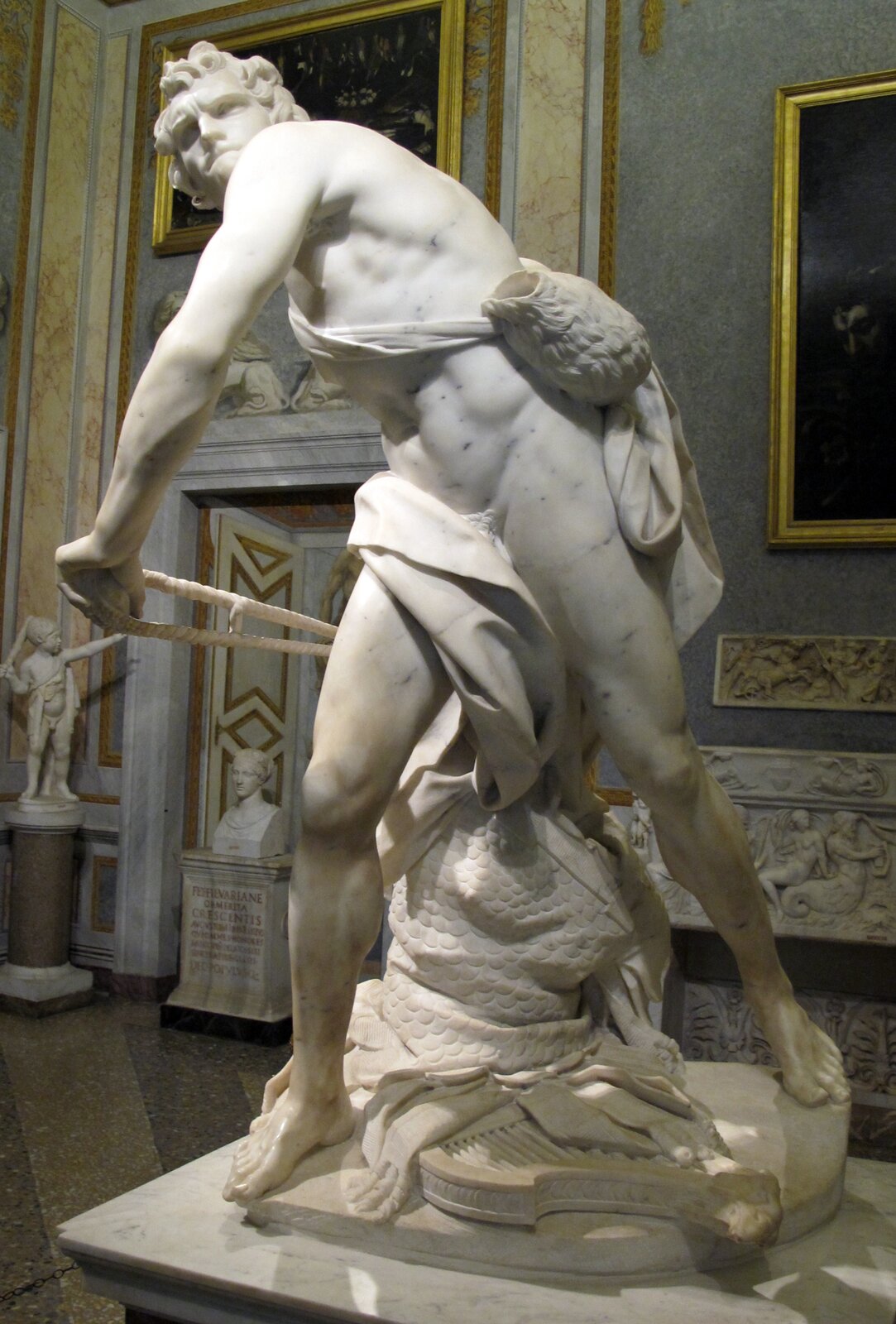 Fotografia rzeźby ukazującej młodego muskularnego mężczyznę o krótkich, kręconych włosach w momencie napinania procy w celu strzelenia z niej. Zaprezentowanie mężczyzny w trakcie tej czynności nadaje dziełu dynamiki. Ciało postaci jest skręcone oraz pochylone. Jego ciało okryte jest szatą przewiązaną przez pas, a przez ramię ma przewieszony worek. Mężczyzna zaciska usta, a jego twarz wyraża skupienie. Pomiędzy jego nogami znajdują się materiały ułożone na fragmencie liry. Dzieło ustawione zostało w jasnym pomieszczeniu o ścianach pokrytych ozdobnymi rzeźbieniami oraz obrazami.