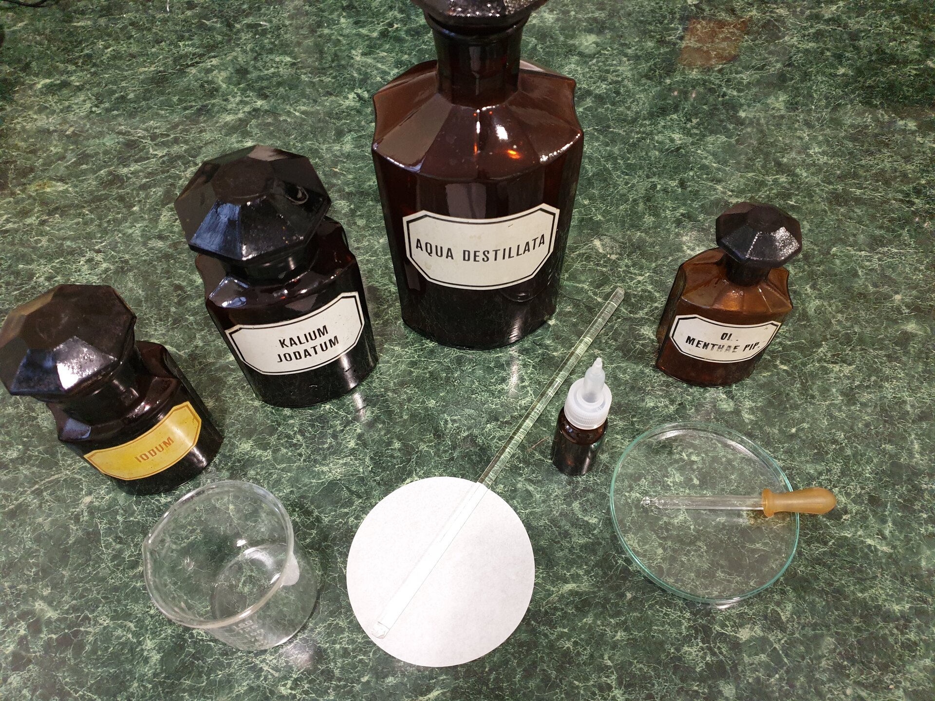 Rycina 2 zatytułowana Krople przedstawia stojące obok siebie szklane butelki ze szlifem, czyli ze szklanym dopasowanym zamknięciem w formie stożkowatej. Butelki mają różne rozmiary. Każda posiada podpisaną etykietę, między innymi: aqua destilata, czyli woda destylowana, kalium jodatum, czyli jodek sodu oraz sodium, czyli soda. Obok stoi niewielka butelka z końcówką dozującą krople, szalka, czyli płaski walcowaty odkryty pojemnik ze szkła. W szalce leży niewielka pipeta. Obok znajduje się niewielka zlewka, sączek oraz szklane mieszadełko w postaci patyczka.