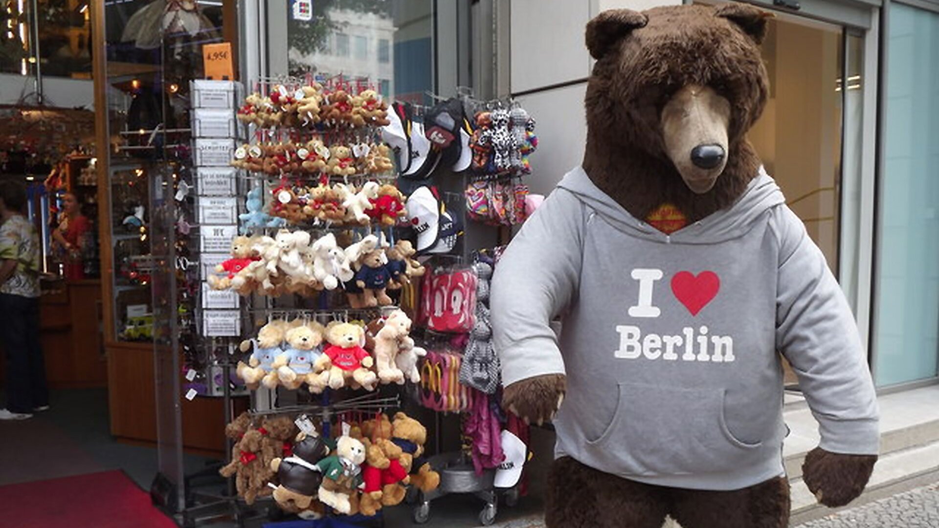 Zdjęcie przedstawia stojaki z pamiątkami, które zostały wystawione na zewnątrz, przed sklep. Na stojakach wiszą różnej wielkości pluszowe misie, czapki z daszkiem, klapki, opaski, magnesy. Na prawo od stojaków, na chodniku widoczny jest stojący pluszowy niedźwiedź brunatny wielkości człowieka, ubrany w szarą bluzę z kapturem z napisem w języku angielskim: I love Berlin. W miejscu słowa love widoczne jest czerwone serduszko. W głębi po lewej stronie widoczne jest wnętrze sklepu.