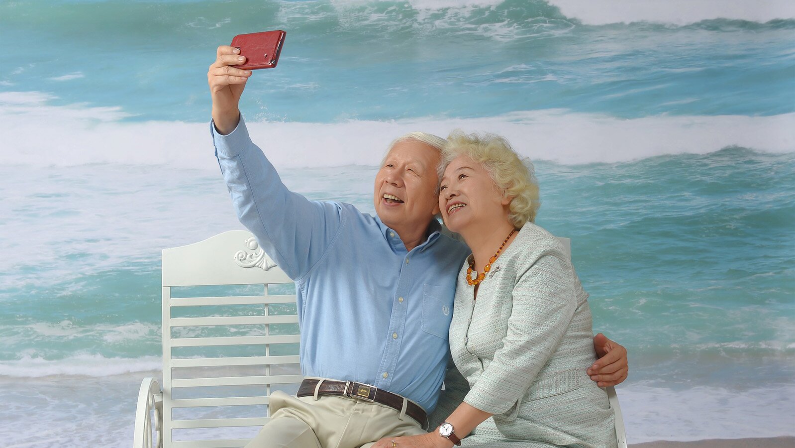 Zdjęcie przedstawia starszego mężczyznę i kobietę, którzy siedzą na białej ławce, nad brzegiem morza. Mężczyzna, siedzący po lewej stronie, uśmiecha się i podnosi prawą rękę do góry. W ręce trzyma telefon, którym próbuje zrobić zdjęcie. Lewą ręką obejmuje kobietę w pasie. Kobieta, siedząca po prawej stronie, uśmiecha się, a jej głowa oparta jest o głowę mężczyzny. Jej lewa ręka leży na kolanie mężczyzny. Z tyłu widoczna jest niebieska woda i fale.