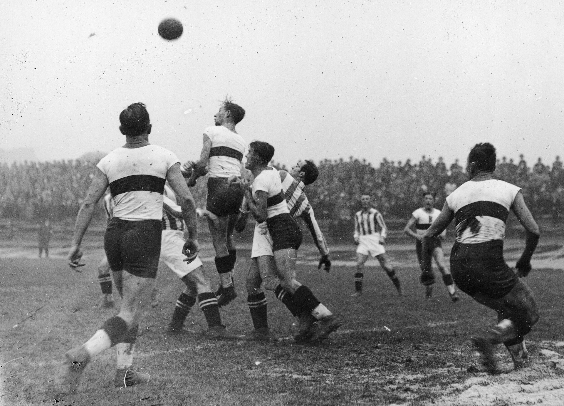 Czarno białe zdjęcie przedstawia mężczyzn grających na boisku w piłkę nożną. Jeden z nich wyskakuje wysoko, aby odbić piłkę głową.