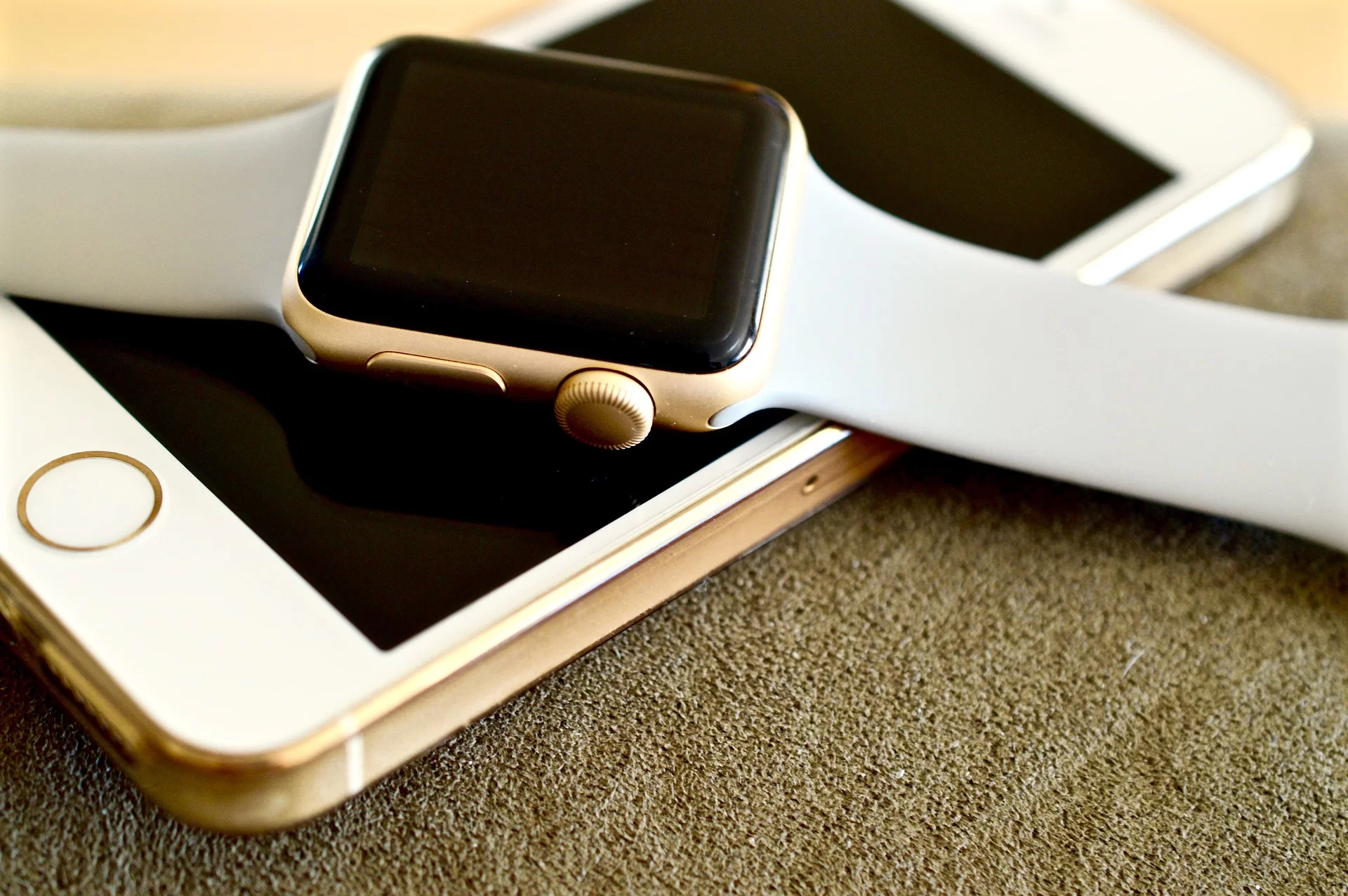 Kliknij, aby powiększyć Na zdjęciu, na podłożu leży apple iPhone. Ma wygaszony ekran. Na nim leży smartwatch, również ma wygaszony ekran. Smartwatch ma kwadratowy ekran i biały pasek.