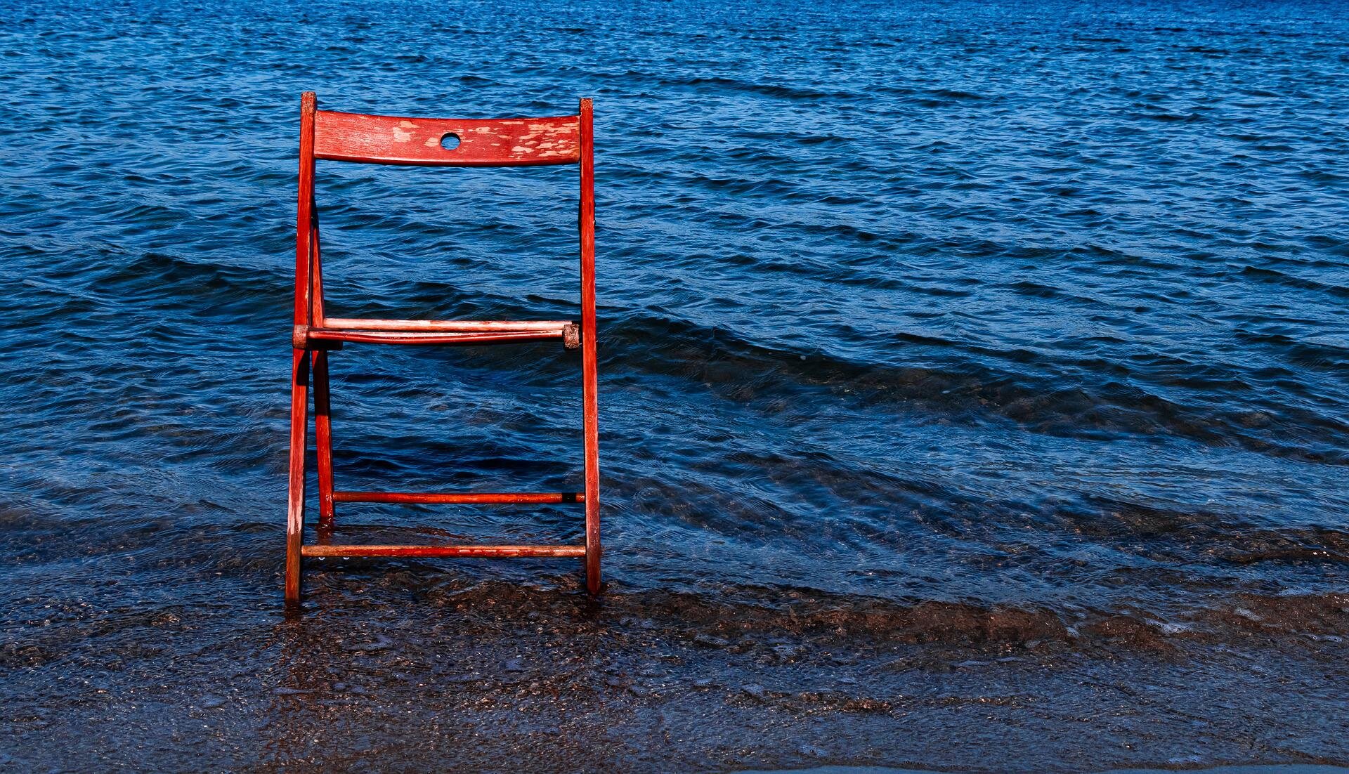 Zdjęcie przedstawia czerwone, drewniane krzesło, które stoi na brzegu morza. Dół krzesła jest częściowo zanurzony w wodzie. Woda jest spokojna, widoczne są niskie fale.