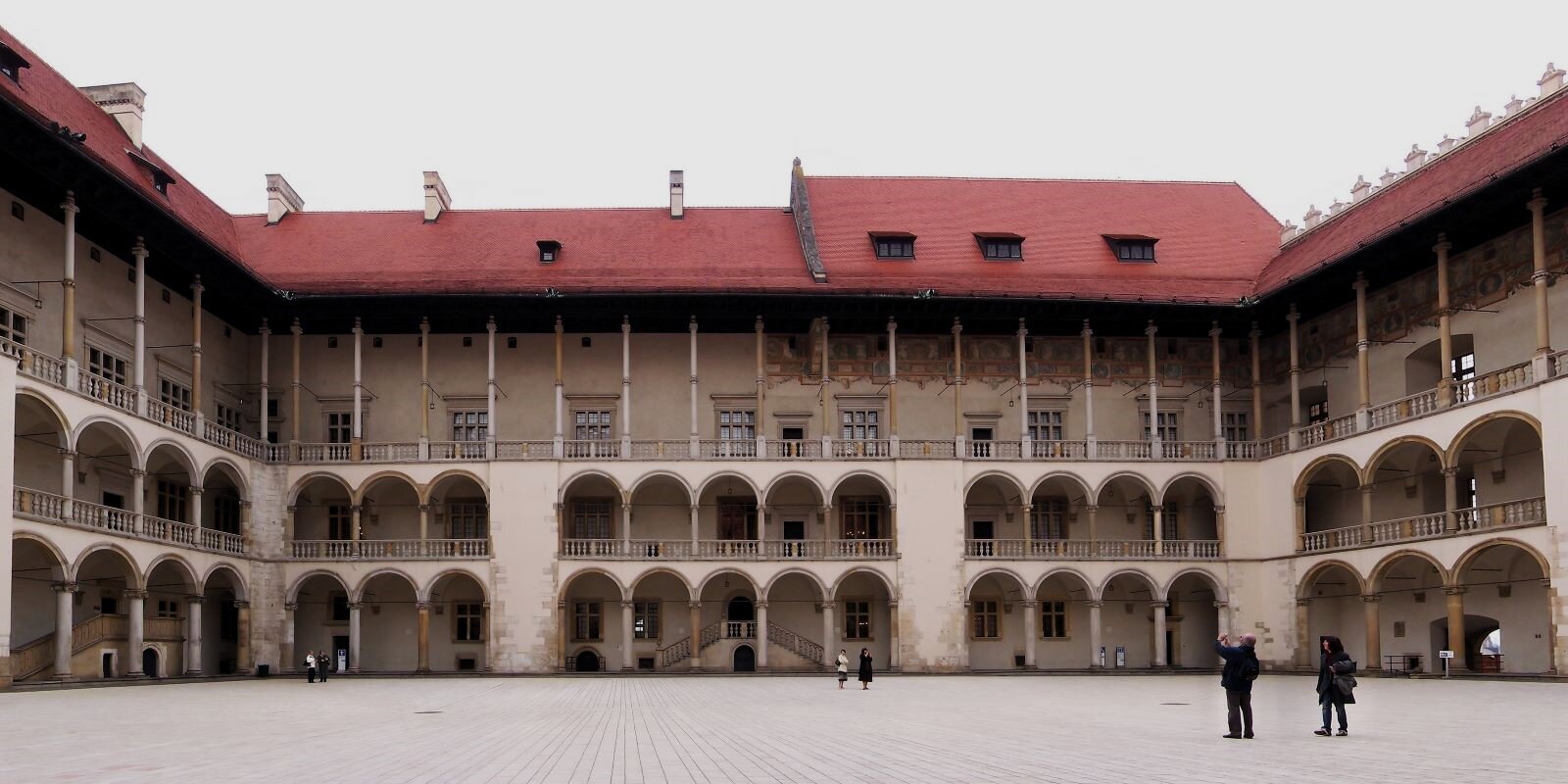 Zdjęcie ukazuje wewnętrzny dziedziniec zamku na Wawelu w Krakowie w kształcie litery U. Jego ozdobą są rytmiczne krużganki zakończone półokrągłymi łukami na dwóch pierwszych kondygnacjach. Dach jest wsparty na prostych, długich kolumnach na ostatniej kondygnacji. Ta konstrukcja dodaje lekkości całej bryle budynku.