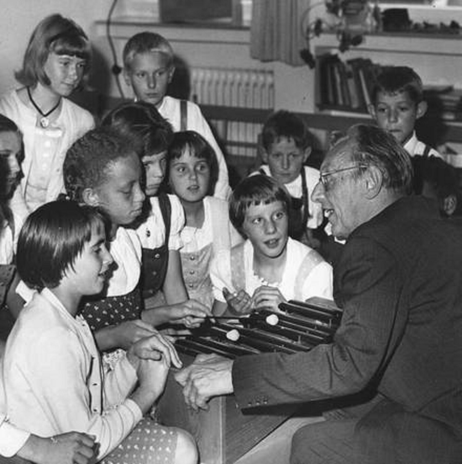 Ilustracja przedstawia Carl'a Orff'a w dziecięcej wiosce pomocy w Dießen. Starszy mężczyzna po prawej stronie jest skupiony na rozmowie z dziećmi, ubrany w ciemny garnitur, nosił okulary. Dzieci w wieku szkolnym wsłuchują się w wypowiedzi Orffe.