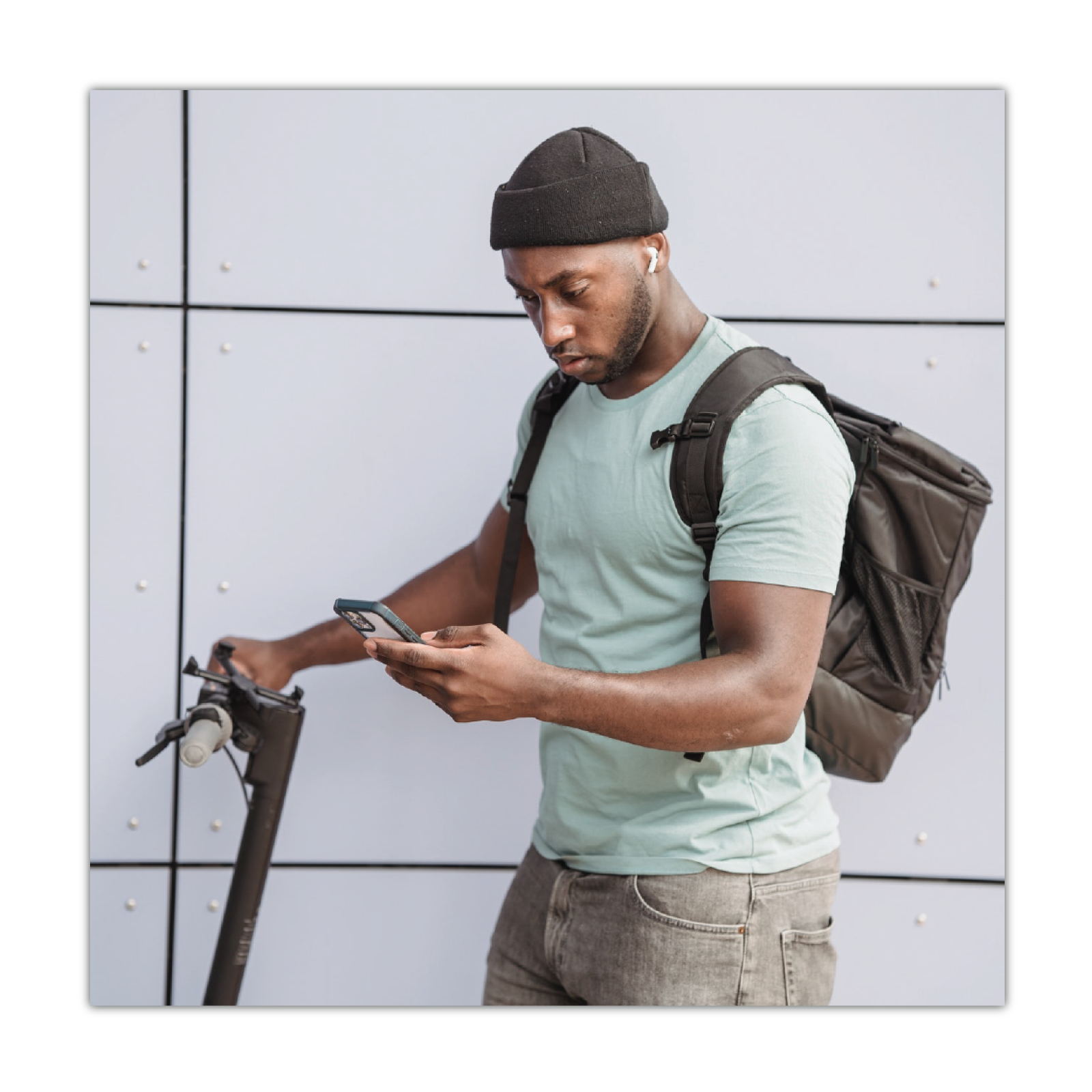 Zdjęcie przedstawia młodego mężczyznę z plecakiem, który prawą dłonią trzyma kierownicę hulajnogi a w lewej ma telefon komórkowy, na który patrzy.