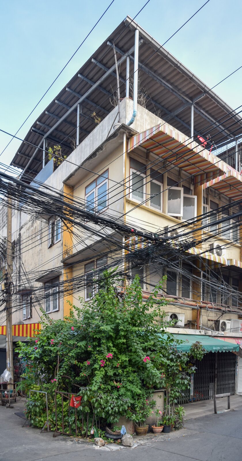 Zdjęcie przedstawia róg wysokiego budynku. Przed nim, nad ulicą, krzyżują się pasy okablowania. Kabli jest bardzo dużo i są poplątane. Niektóre zwisają łukiem nad chodnikami. Widoczna gęsta zieleń przed budynkiem.