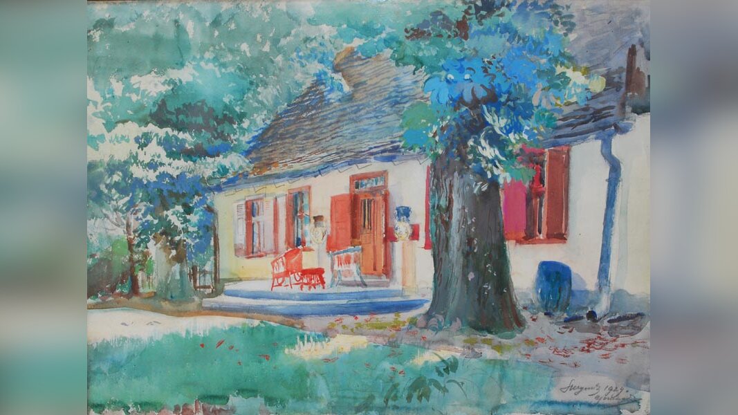 Na obrazie parterowy budynek. Ma białe ściany i ciemny dach. Okna oraz drzwi są pomalowane na czerwono. Przed drzwiami stoi ławka. Wokół budynku rosną drzewa.