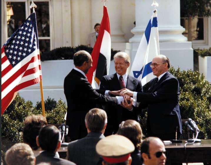 Na zdjęciu znajdują się trzej mężczyźni w garniturach. Jeden z nich jest ciemnoskóry. Za nimi wiszą trzy flagi: Egiptu, Izraela oraz USA. W tle widoczny jest też budynek z białymi filarami oraz zielone krzewy. Na dole fotografii jest kilka osób odwróconych do obiektywu tyłem, widać tylko ich głowy.