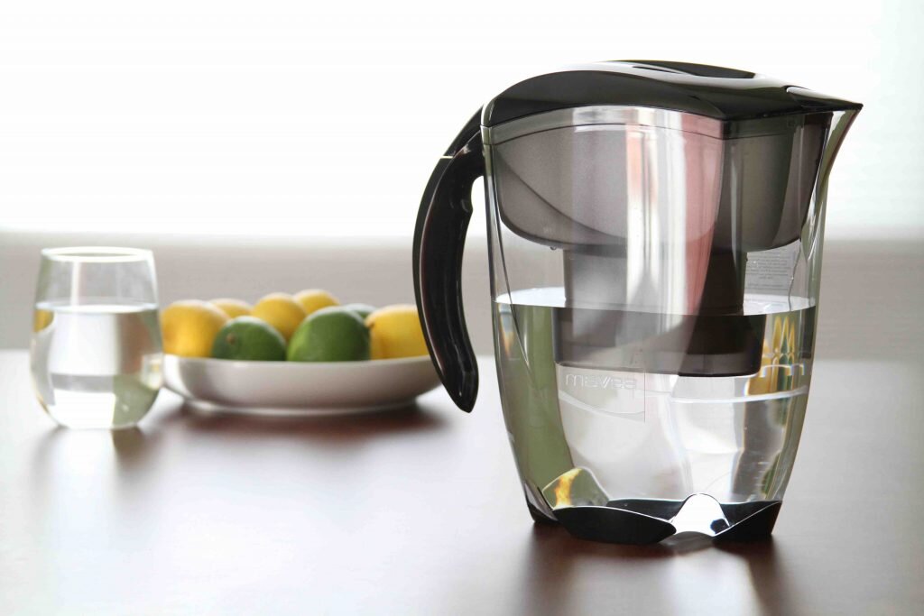 Zdjęcie przedstawia blat na którym stoją: szklanka z wodą, talerz z cytrynami i limonkami, a po prawej stronie dzbanek filtrujący wodę.