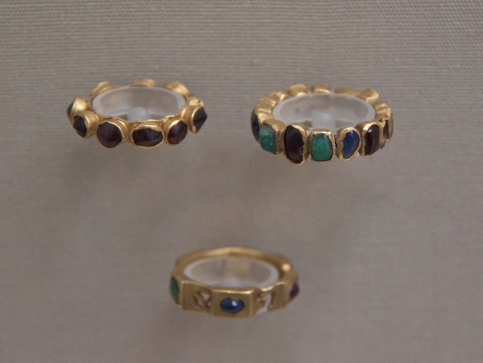 Ilustracja przedstawia zdjęcie trzech złotych pierścieni wysadzanych drogocennymi kamieniami. Kamienie są w kolorach: zielonym, niebieskim, brązowym, czarnym.