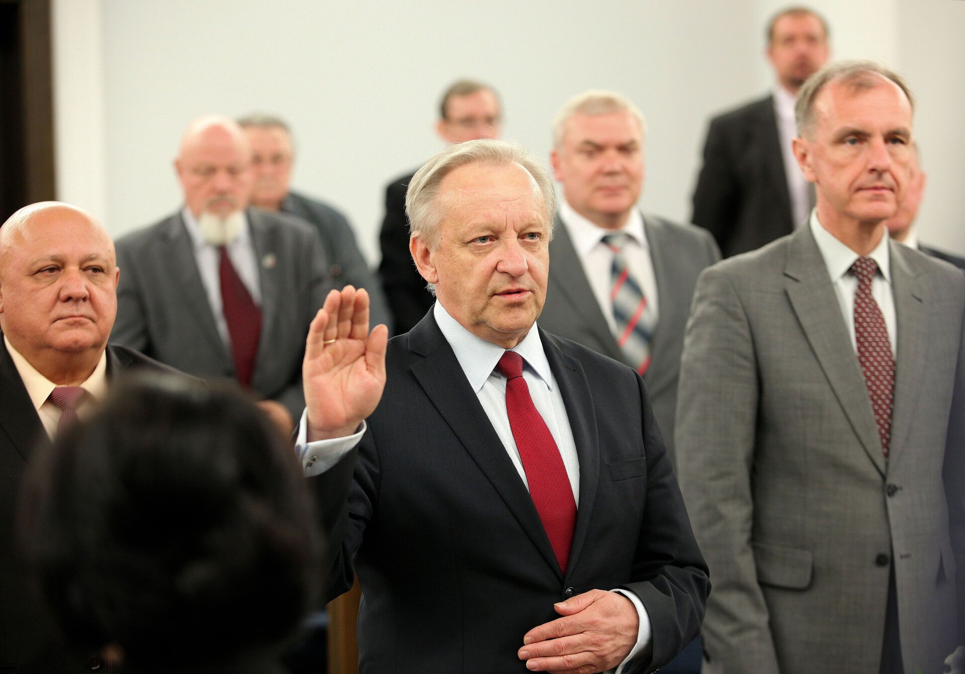 Zdjęcie wykonane w sali senackiej polskiego parlamentu podczas uroczystości. Na pierwszym planie mężczyzna podnosi prawą dłoń otwartą ku górze, zwrócony jest wzrokiem i sylwetką w stronę prawej krawędzi fotografii pod kątem. Po obu jego stronach dwaj mężczyźni w postawie stojącej, za plecami pierwszego rzędu widoczne sylwetki pięciu pozostałych uczestników zgromadzenia. Na pierwszym planie, bliżej lewej dolnej krawędzi widoczny tył głowy postaci, najpewniej pracownika parlamentu. Wszystkie osoby ukazane na fotografii ubrane w stroje galowe - garnitury z krawatami.