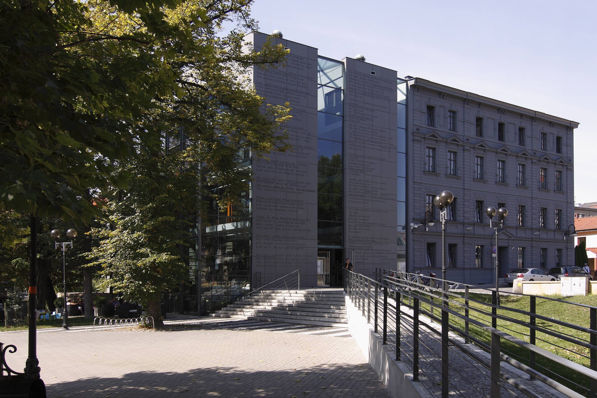 Zdjęcie przedstawia trzypiętrowy szary budynek. Po środku ściany budynku znajduje się przeszklenie. Po prawej stronie część fasady z licznymi oknami. Przed budynkiem, po lewej stronie znajdują się drzewa. 