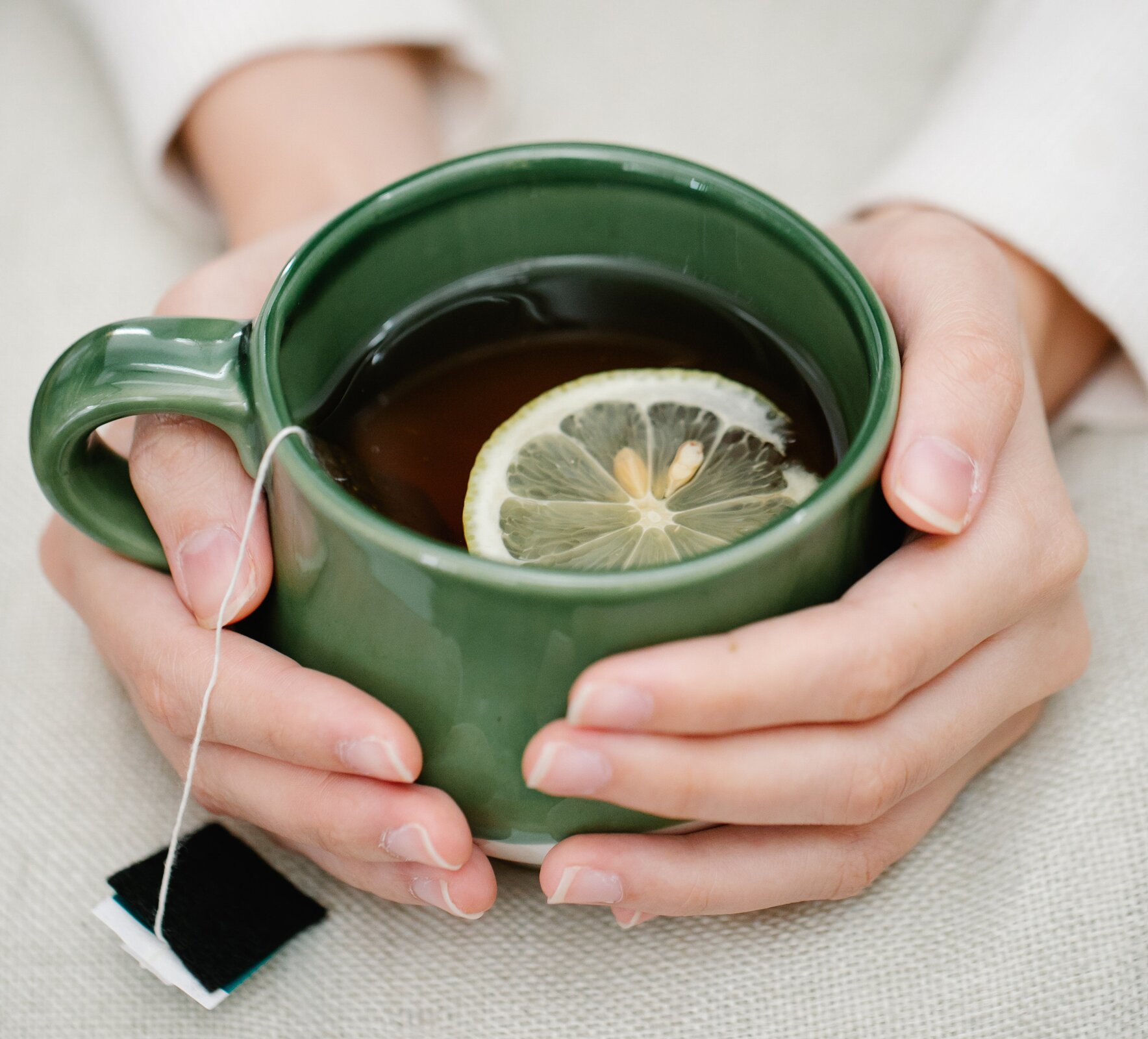 Zdjęcie przedstawia zielony kubek z herbatą i cytryną, który jest trzymany dwoma rękoma przez jakąś osobę. 