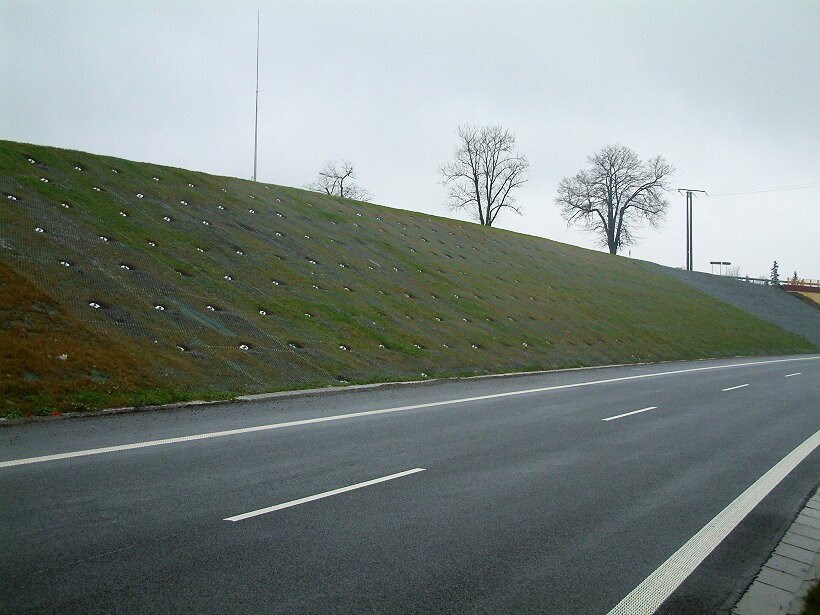 Zdjęcie przedstawia zbocze pagórka biegnące wzdłuż asfaltowej drogi. Zbocze pokryte jest zieloną tkaniną oraz dodatkowo dużymi gwoździami. Gwoździe te powbijane są w zbocze w równomiernych odstępach przytrzymując tkaninę w miejscu i zabezpieczając ziemię przed obsuwaniem się na drogę.