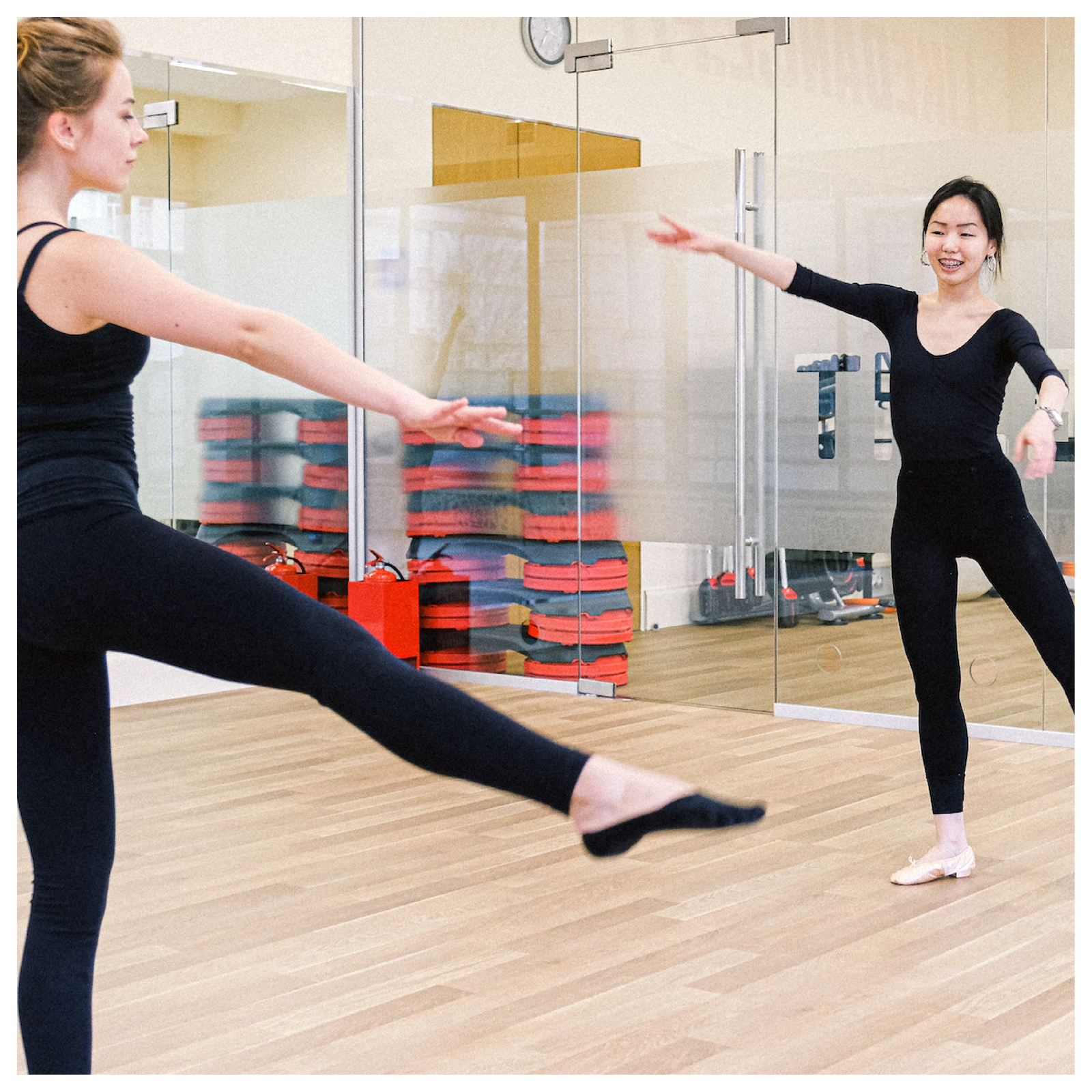 Zdjęcie przedstawia dwie kobiety w sali gimnastycznej, które naprzeciwko siebie ćwiczą balet.