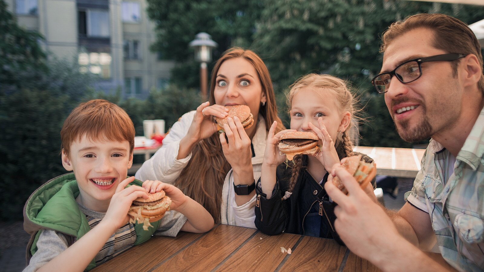 Zdjęcie przedstawia czwórkę ludzi, dwójkę dorosłych i dwójkę dzieci. Wszyscy siedzą przy stole i jedzą hamburgery.