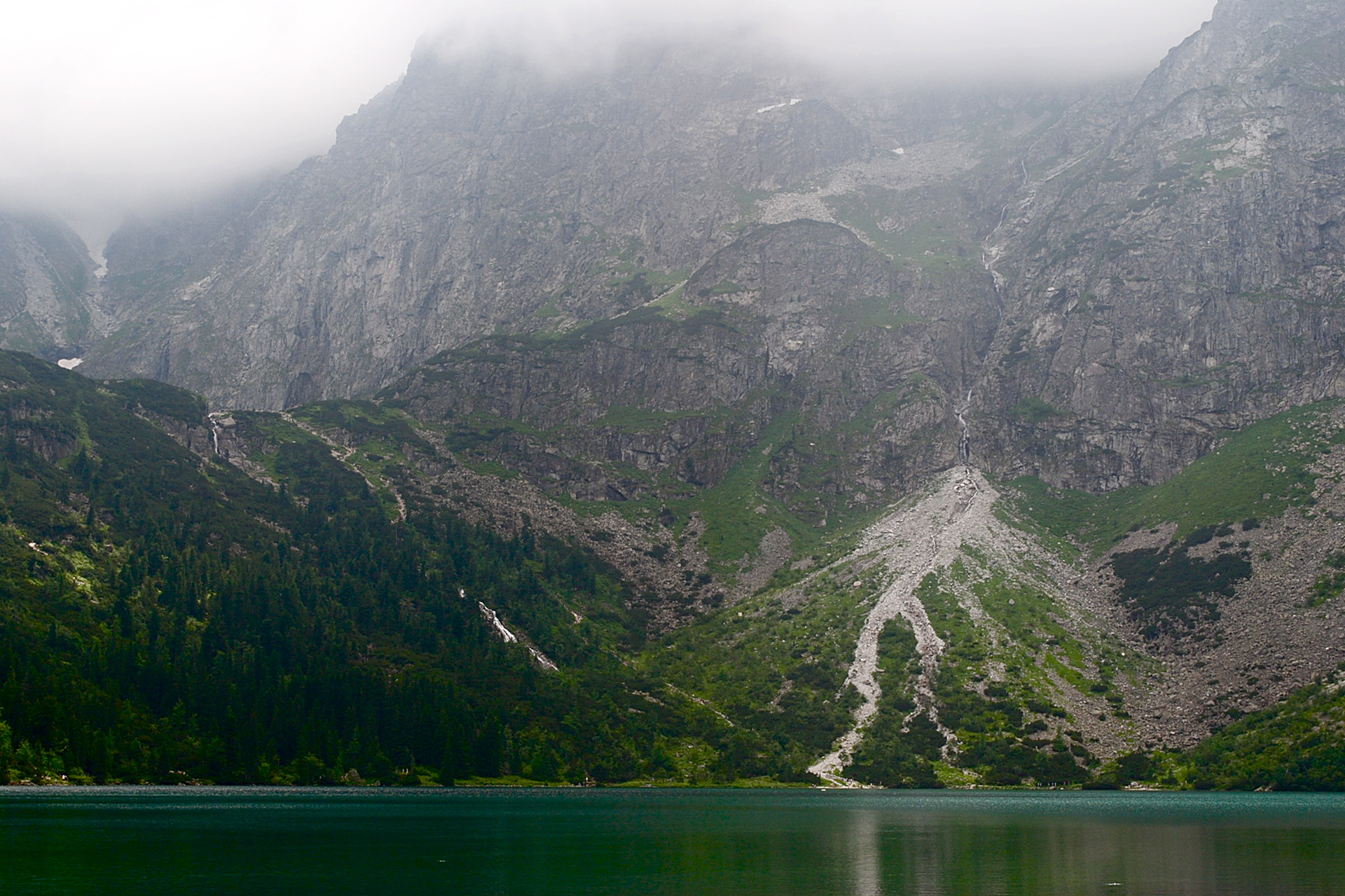 Fotografia prezentuje wysokie, szare góry, porośnięte niewielkimi roślinami i powstałe na nich piargi, czyli jasne, strome usypiska fragmentów skalnych, powstałe u podnóża góry znajdującej się nad jasnym, zielonym, rozległym Morskim Okiem.